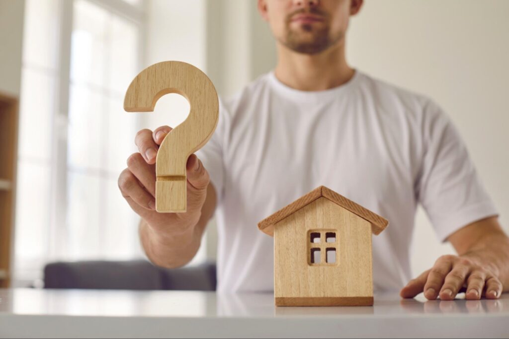 Man holding a question mark next to a wooden house, representing the question, “Why is my house not selling?